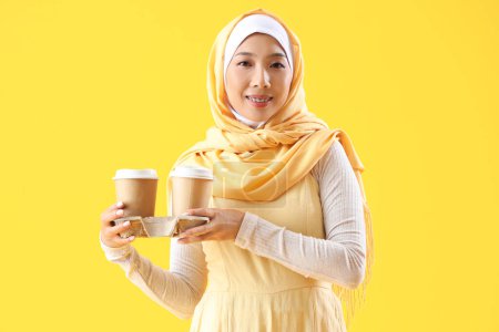 Téléchargez les photos : Belle jeune femme asiatique en hijab avec des tasses de café sur fond jaune - en image libre de droit