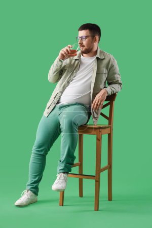 Photo for Young sommelier tasting wine while sitting on wooden chair against green background - Royalty Free Image