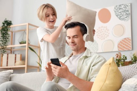 Photo for Young man in headphones using mobile phone and his naughty little son with pillow at home - Royalty Free Image