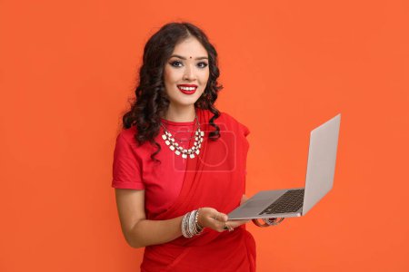 Téléchargez les photos : Belle femme indienne en sari avec ordinateur portable sur fond orange - en image libre de droit