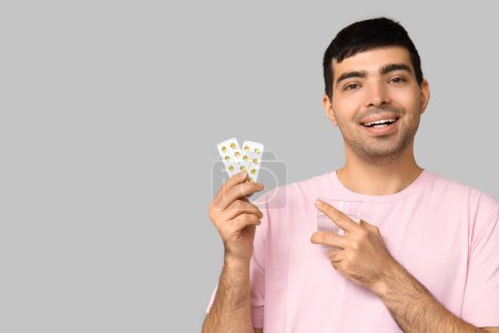 Young man with water pointing at blisters of vitamin A pills on light background