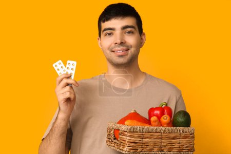 Young man with blisters of vitamin A pills and healthy food on yellow background
