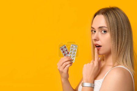 Nachdenkliche junge Frau mit Blasen von Vitamin-A-Pillen auf gelbem Hintergrund, Nahaufnahme