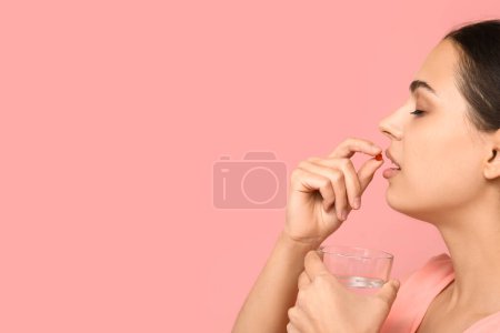 Pretty young woman taking vitamin A capsule on pink background