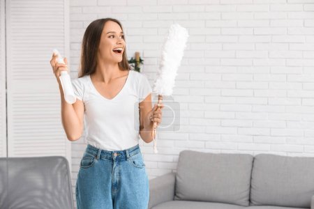Cheerful young woman with pp-duster and bottle of detergent in living room
