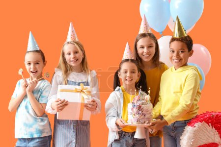 Foto de Lindos niños pequeños en sombreros de fiesta con pastel de cumpleaños y regalos sobre fondo naranja - Imagen libre de derechos