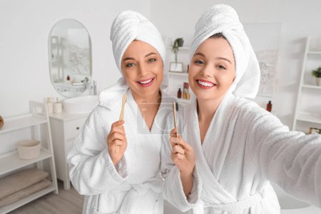 Téléchargez les photos : Jeune femme et sa mère avec des brosses à dents prenant selfie après la douche dans la salle de bain - en image libre de droit