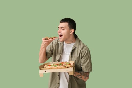 Photo for Young man eating piece of tasty pizza and holding cardboard box on green background - Royalty Free Image