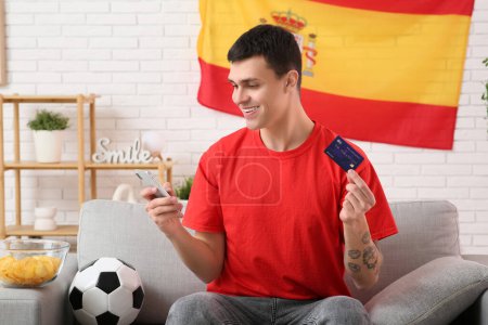 Young man with credit card, mobile phone, soccer ball and flag of Spain on sofa at home