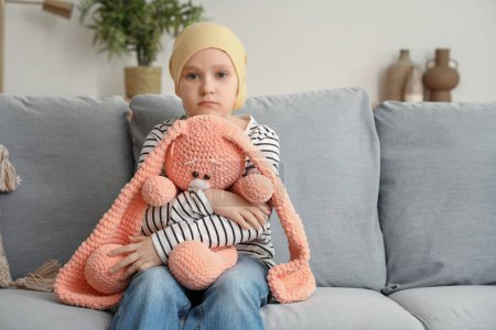 Téléchargez les photos : Triste petite fille après la chimiothérapie avec lapin jouet assis sur le canapé à la maison. Journée internationale du cancer chez les enfants - en image libre de droit