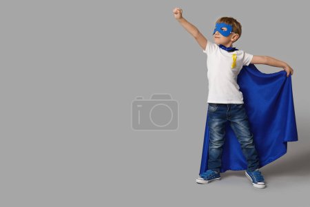 Téléchargez les photos : Mignon petit garçon en costume de super héros avec ruban jaune sur fond gris. Journée internationale du cancer chez les enfants - en image libre de droit