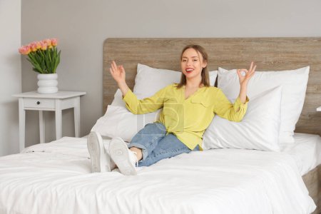 Hübsche junge Frau liegt auf bequemem Bett und zeigt OK im Schlafzimmer