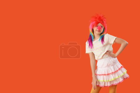 Foto de Chica divertida con peluca colorida y nariz de payaso sobre fondo naranja. Celebración del Día de los Inocentes - Imagen libre de derechos