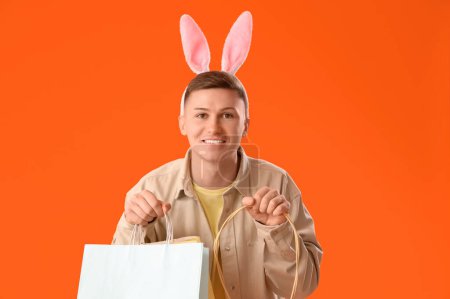 Téléchargez les photos : Jeune homme dans des oreilles de lapin avec des sacs à provisions et panier sur fond orange. Vente de Pâques - en image libre de droit