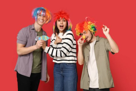 Foto de Jóvenes amigos sorprendidos en disfraz divertido con peces de papel sobre fondo rojo. Celebración del día del tonto de abril - Imagen libre de derechos