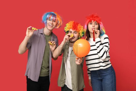 Foto de Jóvenes amigos disfrazados con silbato de fiesta y globo sobre fondo rojo. Celebración del día del tonto de abril - Imagen libre de derechos