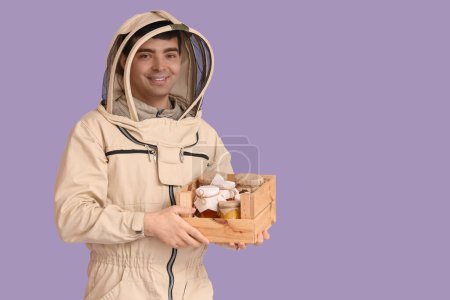 Male beekeeper with box of honey on lilac background