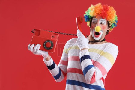Foto de Retrato de payaso sorprendido con teléfono retro sobre fondo rojo. Celebración del día del tonto de abril - Imagen libre de derechos