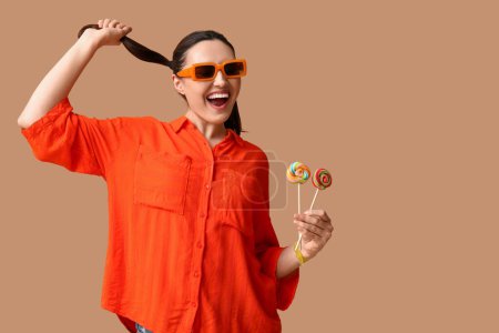 Foto de Hermosa mujer joven en gafas de sol con piruletas dulces sobre fondo marrón - Imagen libre de derechos