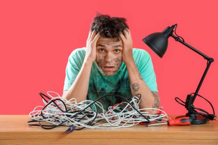 Electrocuted young man with burnt face and wires at table on red background
