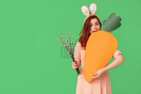 Téléchargez les photos : Beautiful young woman in bunny ears with pussy willow branches and paper carrot on green background. Easter celebration - en image libre de droit