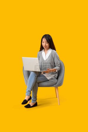 Téléchargez les photos : Belle femme asiatique utilisant un ordinateur portable en fauteuil gris sur fond jaune - en image libre de droit