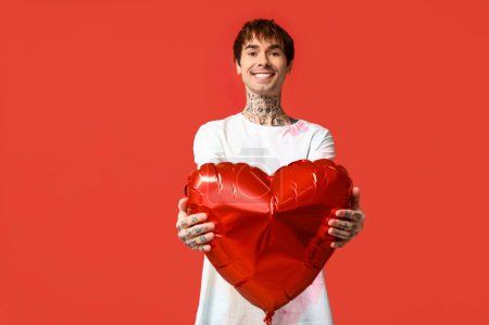 Handsome young man with heart-shaped air balloon on red background
