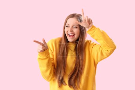 Laughing young woman showing loser gesture and pointing at something on pink background