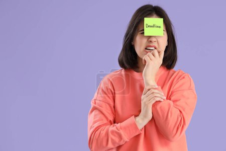 Beautiful young stressed woman holding sticky note with word DEADLINE on purple background. Stress Awareness Month