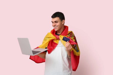 Jeune homme avec drapeau de l'Espagne, carte de crédit et ordinateur portable moderne sur fond rose