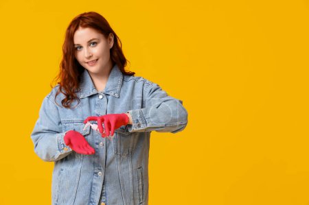 Schöne junge glückliche Frau in Gummihandschuhen, die Desinfektionsmittel auf gelbem Hintergrund anwendet