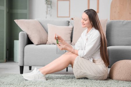 Young woman with bottle of CBD oil sitting on floor at home