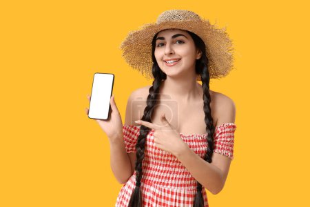 Foto de Joven mujer feliz señalando el teléfono móvil sobre fondo amarillo. Festa Junina (Festival de Junio) celebración - Imagen libre de derechos