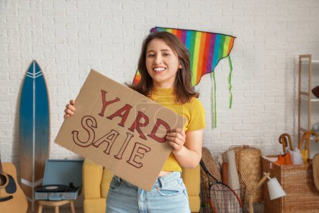 Photo for Young woman holding cardboard with text YARD SALE in room of unwanted stuff - Royalty Free Image