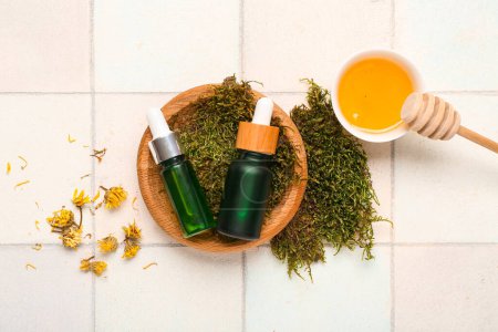 Bottles of essential oil with honey, dried dandelion and green moss on light tile background