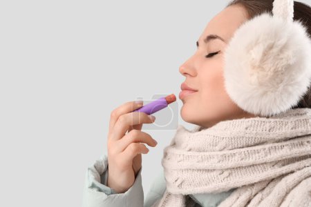 Mujer joven en ropa de invierno aplicando bálsamo labial sobre fondo claro, primer plano
