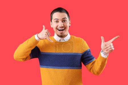 Happy young man showing "call me" gesture and pointing at something on red background