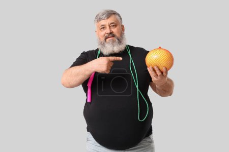 Übergewichtiger älterer Mann mit Springseil, das auf Pomelofrüchte vor grauem Hintergrund zeigt. Konzept zur Gewichtsreduktion