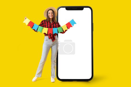 Beautiful young happy woman with garland and big smartphone on yellow background. Festa Junina (June Festival) celebration