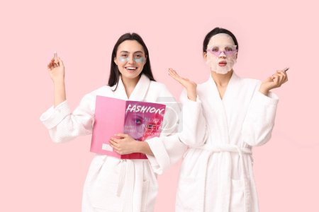 Young women in bathrobes with facial mask, under-eye patches, lip balms and magazine on pink background