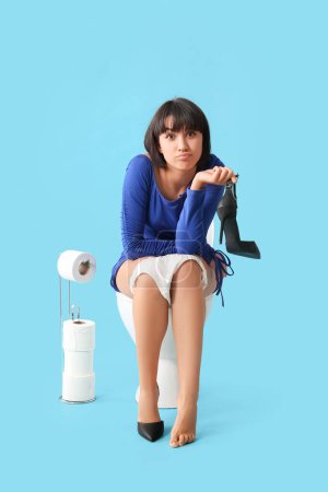 Téléchargez les photos : Jeune femme avec chaussure assise sur un bol de toilette sur fond bleu - en image libre de droit