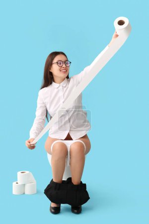 Téléchargez les photos : Jeune femme d'affaires asiatique avec rouleau de papier assis sur un bol de toilette sur fond bleu - en image libre de droit