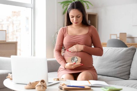 Young pregnant woman with piggy bank sitting on sofa at home. Maternity Benefit concept