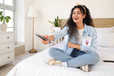 Téléchargez les photos : Belle jeune femme afro-américaine avec panier de crème glacée et télécommande TV assis sur le lit à la maison - en image libre de droit