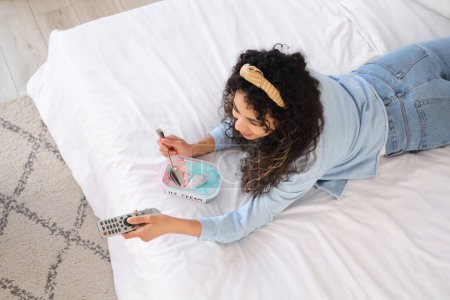Téléchargez les photos : Belle jeune femme afro-américaine avec panier de crème glacée et télécommande TV couchée sur le lit à la maison - en image libre de droit