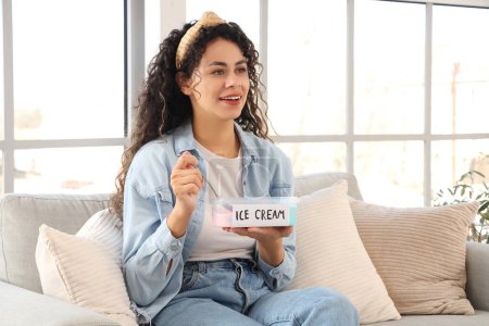 Téléchargez les photos : Belle jeune femme afro-américaine avec panier de crème glacée assis sur le canapé dans le salon - en image libre de droit