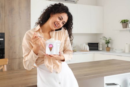 Téléchargez les photos : Belle jeune femme afro-américaine avec panier de crème glacée parlant par téléphone portable dans la cuisine - en image libre de droit