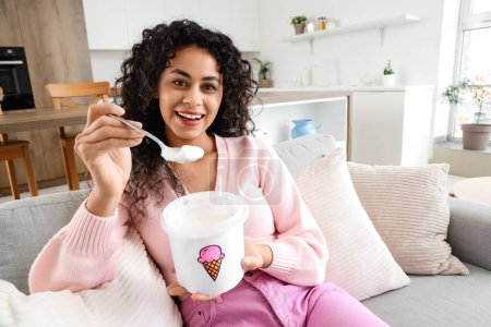 Téléchargez les photos : Belle jeune femme afro-américaine avec panier de crème glacée assis sur le canapé à la maison - en image libre de droit