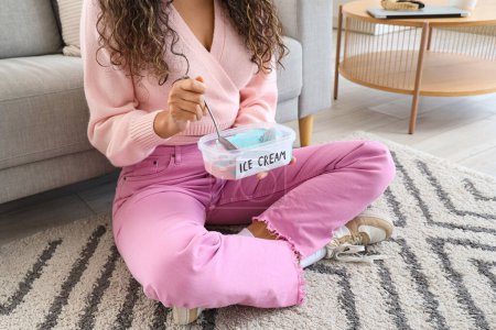 Téléchargez les photos : Jeune femme afro-américaine avec panier de crème glacée assis sur le tapis à la maison - en image libre de droit