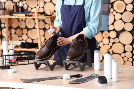 Woman shoemaker applying water repellent spray over pair of leather male shoes at wooden table